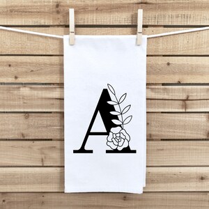 以下が含まれることがあります： 黒いアルファベットのAと黒い花のデザインが施された白いキッチンタオル。Aは太字で、花のデザインは花と葉のシンプルなアウトラインです。
