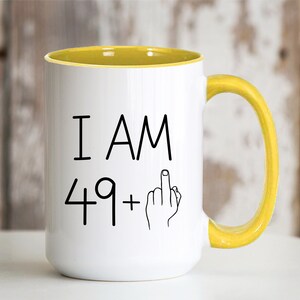 May include: White coffee mug with a yellow rim and handle. The mug has black text that reads "I AM 49+" with a middle finger emoji.