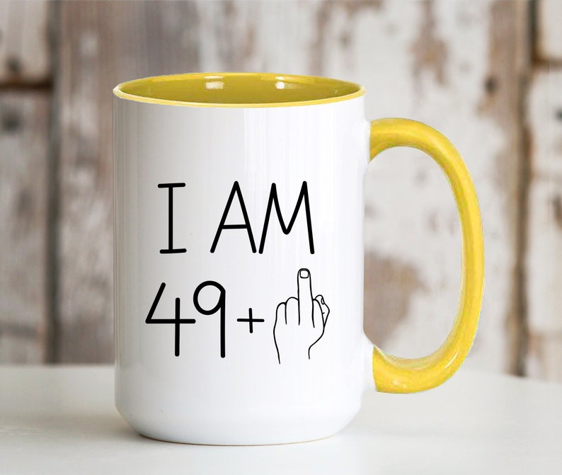 May include: White coffee mug with a yellow rim and handle. The mug has black text that reads "I AM 49+" with a middle finger emoji.