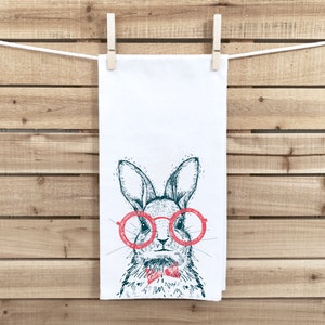 May include: A white kitchen towel with a hand-drawn illustration of a rabbit wearing red-rimmed glasses and a red bow tie.