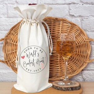 May include: White wine bottle bag with a black circle design that says "Soon to be World's Coolest Aunt Est. 2023".