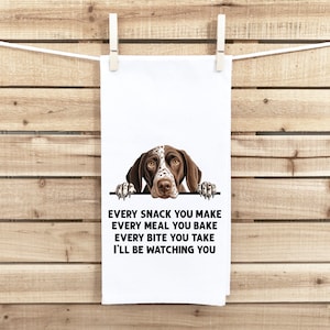 May include: A white kitchen towel with a brown and white German Shorthaired Pointer peeking over the top. The text on the towel reads "Every snack you make, every meal you bake, every bite you take, I'll be watching you."