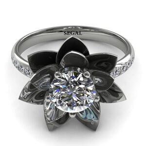 May include: A silver ring featuring a large, clear, round-cut diamond at the center, surrounded by dark gray petals in a floral design. The band is adorned with small, clear stones. The word "SEGAL" is engraved on the ring.
