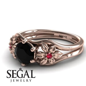 Op de afbeelding: Een verlovingsring van ros&eacute;goud met een zwarte diamant en twee robijn accenten. De ring heeft een bloemmotief met twee delicate bloemen aan weerszijden van de middelste steen. De band is gegraveerd met het woord "SEGAL".