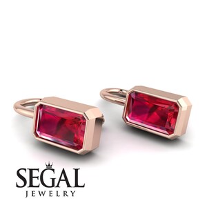 Pode incluir: Brincos em ouro rosa com pedras preciosas vermelhas retangulares. Os brincos têm um design simples e elegante com uma moldura. O logotipo "SEGAL JEWELRY" está na parte inferior.