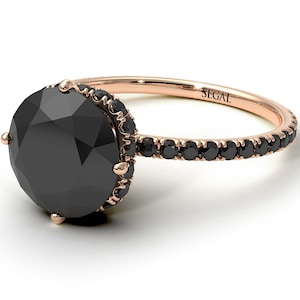 May include: A rose gold engagement ring with a black diamond center stone and a black diamond halo. The band is also set with black diamonds. The ring is engraved with the word "SEGAL".