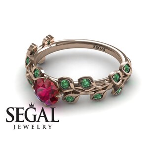 May include: A rose gold ring with a ruby gemstone and emerald green leaves. The ring is engraved with the word "SEGAL".