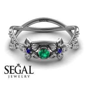 K&ouml;nnte beinhalten: Ein silberner Ring mit einem gr&uuml;nen Smaragd, der von zwei blauen Saphiren umgeben ist. Der Ring hat ein florales Design mit filigranen Details und eine Reihe kleiner Diamanten auf jeder Seite.