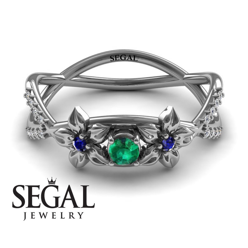 K&ouml;nnte beinhalten: Ein silberner Ring mit einem gr&uuml;nen Smaragd, der von zwei blauen Saphiren umgeben ist. Der Ring hat ein florales Design mit filigranen Details und eine Reihe kleiner Diamanten auf jeder Seite.