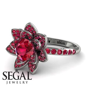 May include: A silver ring with a red ruby center stone and a flower design. The ring is set with smaller red rubies around the center stone and on the band. The ring is engraved with the word "SEGAL".