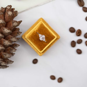 May include: A silver ring with a large oval-shaped diamond in a yellow velvet ring box. The box is on a white surface with coffee beans scattered around it.