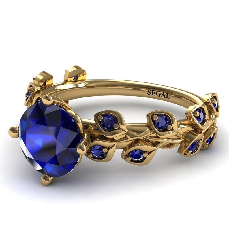 May include: A gold ring with a large blue sapphire gemstone and a delicate leaf design with smaller blue sapphires. The ring is engraved with the word "SEGAL".