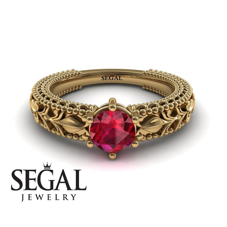 May include: A gold ring with a red ruby gemstone set in a delicate floral design. The band is adorned with small beads and intricate details. The ring is engraved with the word "SEGAL".