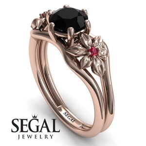 Op de afbeelding: Een verlovingsring van ros&eacute;goud met een zwarte diamant als middelste steen en twee robijn accentstenen. De ring heeft een bloemmotief met delicate details. De band is gegraveerd met het woord "SEGAL".