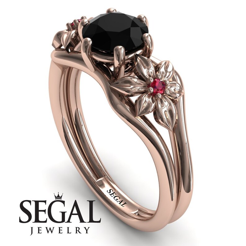 Op de afbeelding: Een verlovingsring van ros&eacute;goud met een zwarte diamant als middelste steen en twee robijn accentstenen. De ring heeft een bloemmotief met delicate details. De band is gegraveerd met het woord "SEGAL".