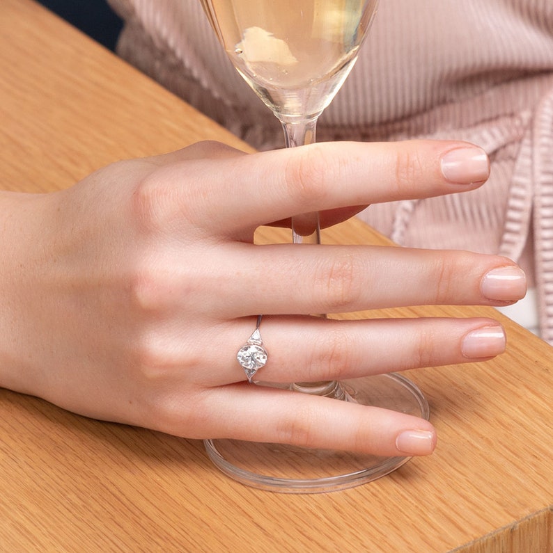 May include: A close-up of a hand wearing a silver ring with a large, round, clear gemstone. The ring has two smaller, clear gemstones on either side of the main stone.