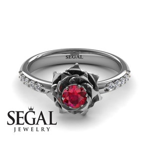 May include: A silver ring with a red ruby set in a rose design. The band is accented with small diamonds. The ring is engraved with the words "SEGAL JEWELRY".