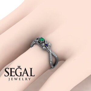 K&ouml;nnte beinhalten: Ein silberner Verlobungsring mit einem gr&uuml;nen Smaragd und zwei blauen Saphiren. Der Ring hat ein florales Design mit Diamant-Akzenten und einem Kreuzband.