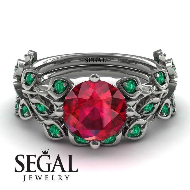 May include: A ruby and emerald engagement ring set with a large red ruby centre stone surrounded by smaller green emeralds. The ring is set in a silver band with a leafy design.