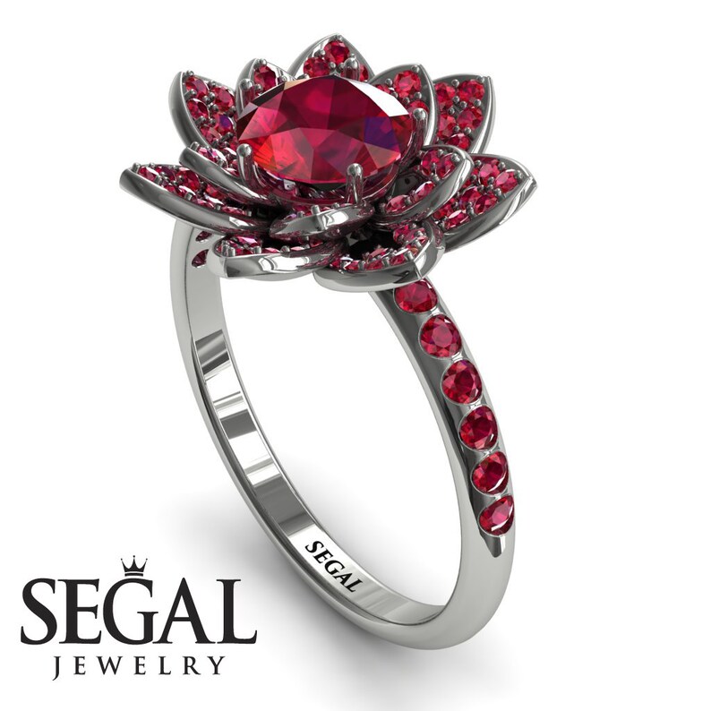 May include: A red ruby gemstone engagement ring set in a white gold band. The ring features a lotus flower design with smaller red rubies surrounding the center stone. The band is engraved with the word "SEGAL".