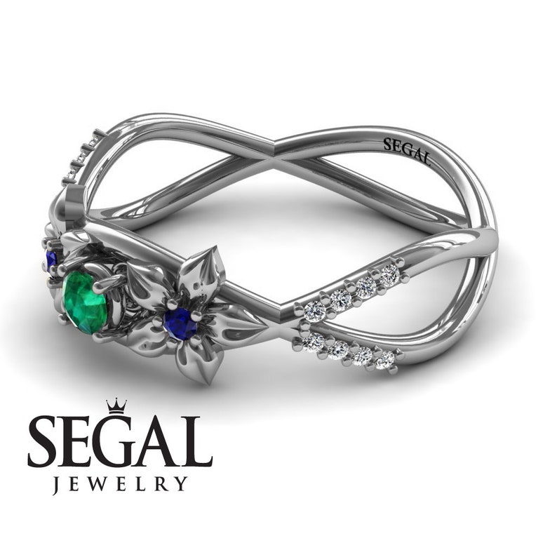 K&ouml;nnte beinhalten: Ein silberner Ring mit einem floralen Design, das mit einem Smaragd und einem Saphir besetzt ist. Der Ring hat ein gekreuztes Band mit einer Reihe kleiner Diamanten.