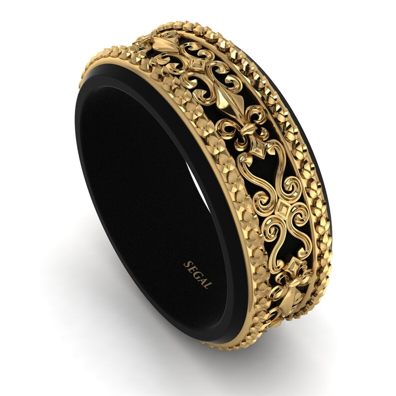 Op de afbeelding: Een zwarte en gouden ring met een ingewikkeld ontwerp. De ring heeft een zwarte band met een gouden band erboven. De gouden band heeft een herhalend patroon van fleur-de-lis en andere decoratieve elementen. De ring is gegraveerd met het woord "SEGAL".