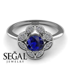 May include: A silver ring with a blue sapphire center stone surrounded by a halo of smaller diamonds. The ring features a floral design with four petals.