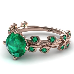 May include: A rose gold ring with a large emerald green gemstone and smaller emerald green gemstones set in a leaf design. The ring is engraved with the word "SEGAL".
