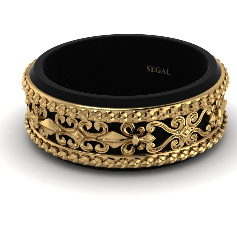 Op de afbeelding: Een zwarte en gouden ring met een decoratief ontwerp. De ring heeft een zwarte band met een gouden band erboven. De gouden band heeft een decoratief ontwerp met een fleur-de-lis-patroon. De ring is gegraveerd met het woord "SEGAL".