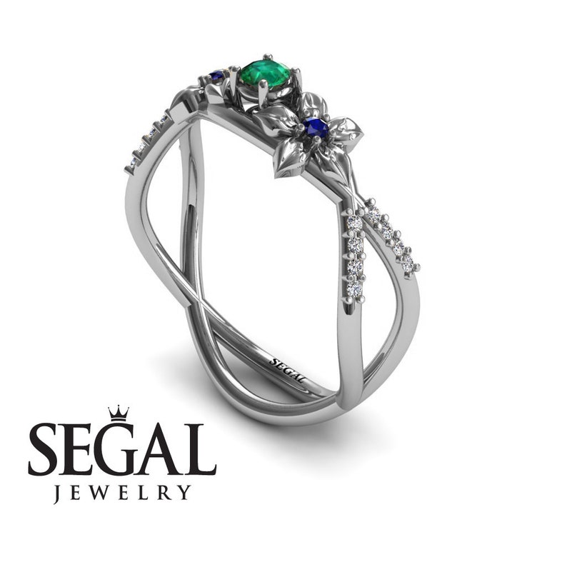 K&ouml;nnte beinhalten: Ein silberner Ring mit einem gr&uuml;nen Smaragd und blauen Saphiren, die in einem floralen Design gefasst sind. Der Ring hat ein Kreuzband mit kleinen Diamanten.