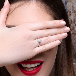 May include: A woman with long brown hair is smiling and covering her eyes with her hand. She is wearing a silver ring with a large diamond on her ring finger.