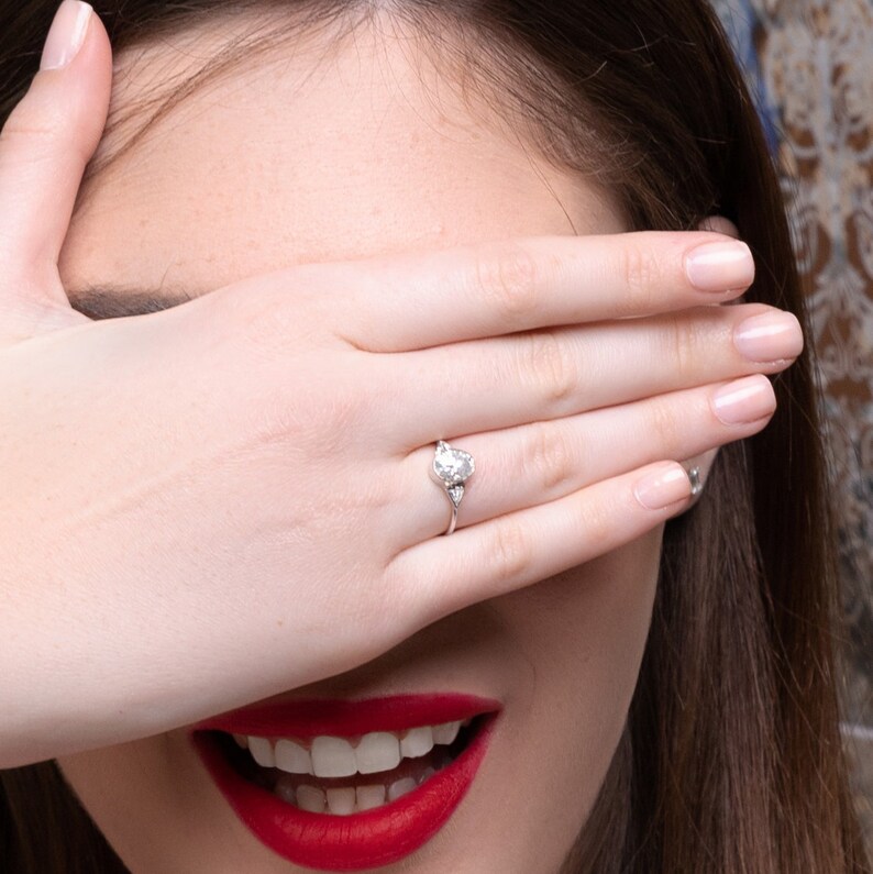 May include: A woman with long brown hair is smiling and covering her eyes with her hand. She is wearing a silver ring with a large diamond on her ring finger.