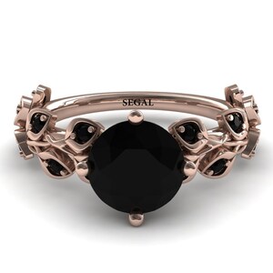 May include: A rose gold ring with a black diamond center stone and black diamond accents on the band. The band is decorated with a leaf design. The ring is engraved with the word "SEGAL".