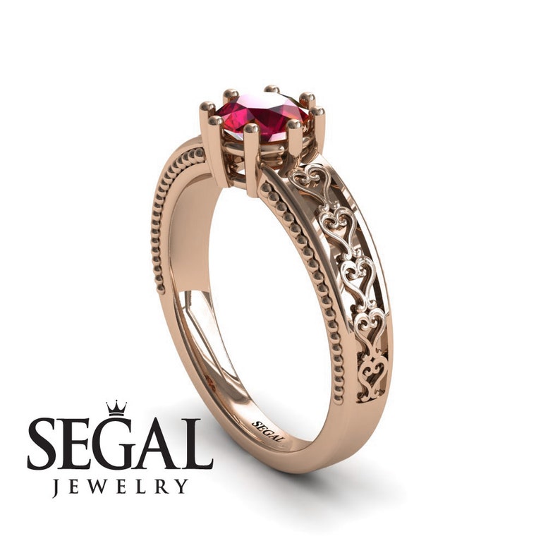 May include: A rose gold engagement ring with a red gemstone and a delicate heart design on the band. The ring is engraved with the word "SEGAL".