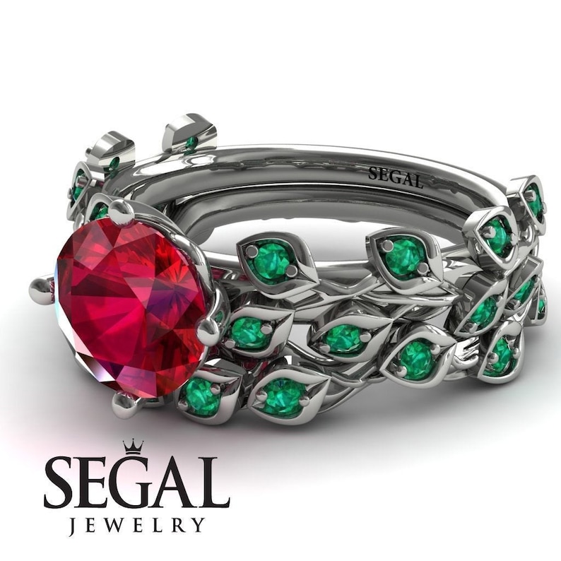 May include: A silver ring set with a large, round, red ruby gemstone surrounded by a band of smaller, emerald green gemstones. The band is designed with a leafy pattern. The ring is engraved with the word "SEGAL".