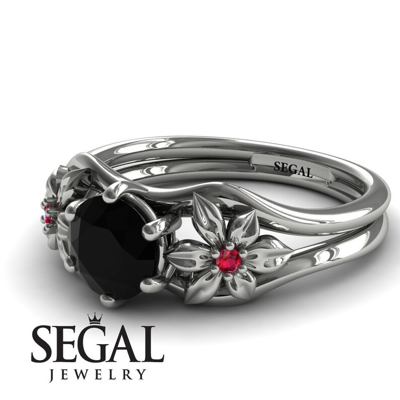 May include: A black diamond engagement ring with two ruby accent stones set in a floral design. The ring is made of white gold and has the inscription "SEGAL" on the band.
