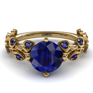 May include: A gold ring with a large blue sapphire center stone and smaller blue sapphires set in leaf-shaped accents. The ring is inscribed with the word "SEGAL".