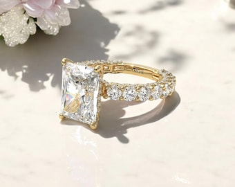 Op de afbeelding: Een gouden verlovingsring met een grote, rechthoekige, smaragd geslepen diamant als middelste steen en een band van kleinere ronde diamanten. De ring ligt op een wit oppervlak en de merknaam "SEGAL" is zichtbaar op de band.