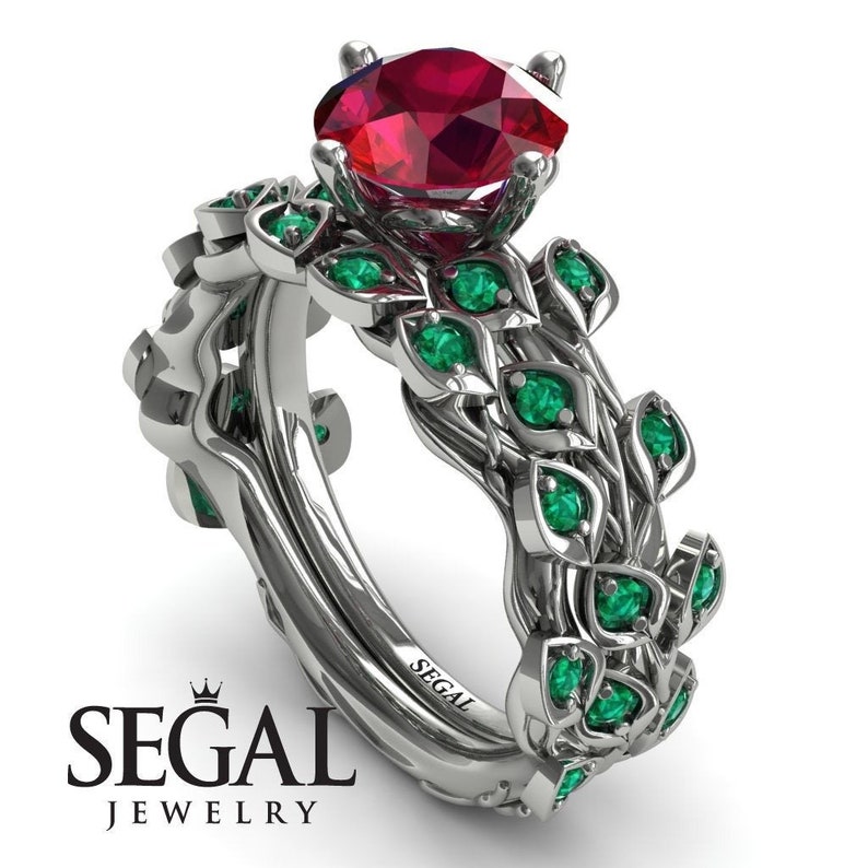 May include: A silver engagement ring set with a large red ruby gemstone and smaller green gemstones set in a leafy design. The ring is engraved with the word "SEGAL".