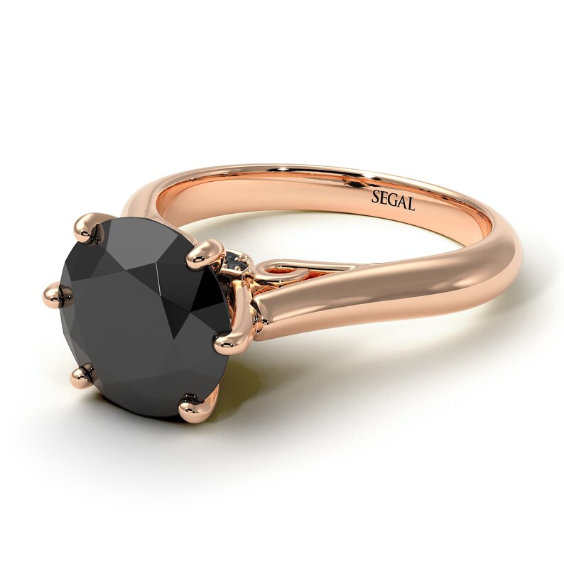 May include: A rose gold engagement ring with a black diamond center stone and a smaller diamond accent. The ring is engraved with the word "SEGAL".