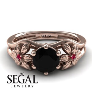 Op de afbeelding: Een verlovingsring van ros&eacute;goud met een zwarte diamant in het midden en twee robijn accentstenen. De ring heeft een bloemmotief met twee delicate bloemen aan weerszijden van de middelste steen. De ring is gegraveerd met het woord "SEGAL".