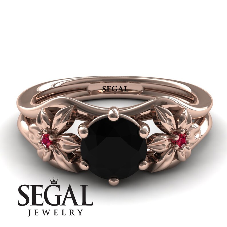 Op de afbeelding: Een verlovingsring van ros&eacute;goud met een zwarte diamant in het midden en twee robijn accentstenen. De ring heeft een bloemmotief met twee delicate bloemen aan weerszijden van de middelste steen. De ring is gegraveerd met het woord "SEGAL".
