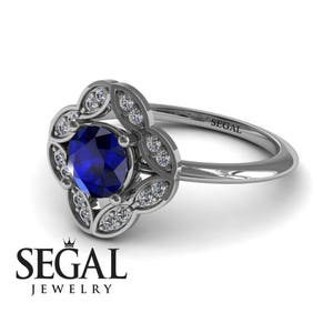 May include: A silver engagement ring with a blue sapphire center stone surrounded by a halo of smaller diamonds. The ring is set in a floral design with a delicate, intricate pattern. The band is simple and sleek, with the inscription "SEGAL" on the inside.