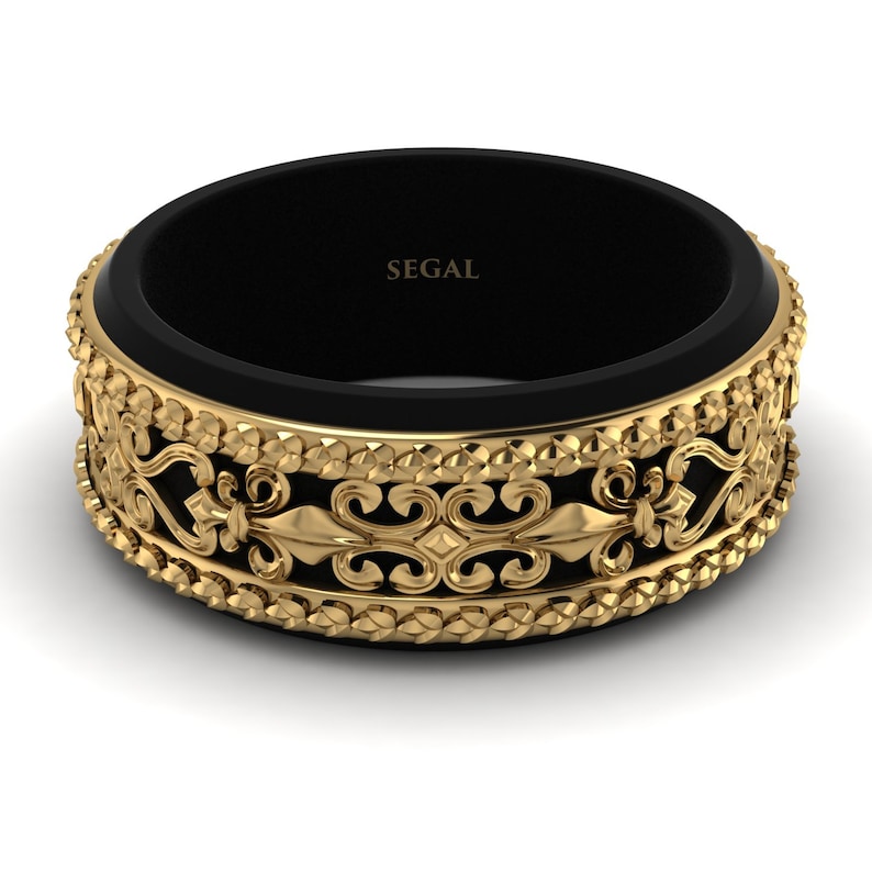 Op de afbeelding: Een zwarte en gouden herenring met een decoratief, versierd ontwerp. De ring heeft een zwarte band met een gouden band aan de buitenrand. De gouden band heeft een herhalend patroon van fleur-de-lis en andere decoratieve elementen. De ring is gegraveerd met het woord "SEGAL".