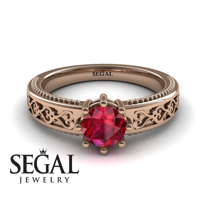 May include: A rose gold engagement ring with a delicate filigree design and a red ruby gemstone. The ring is engraved with the word "SEGAL".