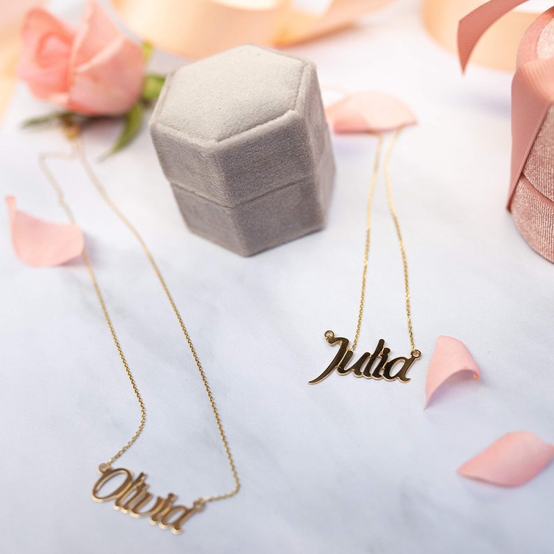 Puede incluir: Dos collares dorados con nombres, uno con "Olivia" y otro con "Julia", sobre una superficie blanca. Una caja de joyer&iacute;a de terciopelo gris y p&eacute;talos de rosa rosas dispersos tambi&eacute;n est&aacute;n presentes. Los collares son delicados y elegantes.