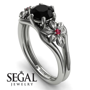 May include: A black diamond engagement ring set with a floral design. The ring features a black diamond center stone and two red ruby accents. The band is made of white gold and has a delicate floral design.