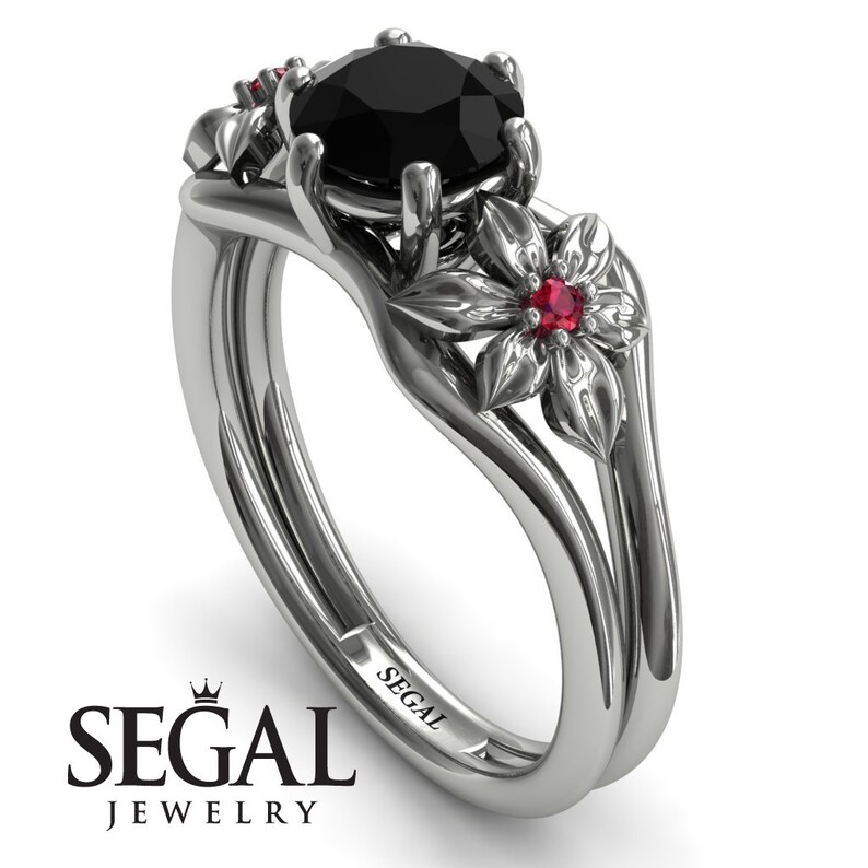 May include: A black diamond engagement ring set with a floral design. The ring features a black diamond center stone and two red ruby accents. The band is made of white gold and has a delicate floral design.