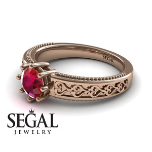 May include: A rose gold engagement ring with a red ruby gemstone and a delicate filigree design. The band is adorned with a beaded edge. The ring is engraved with the word "SEGAL".