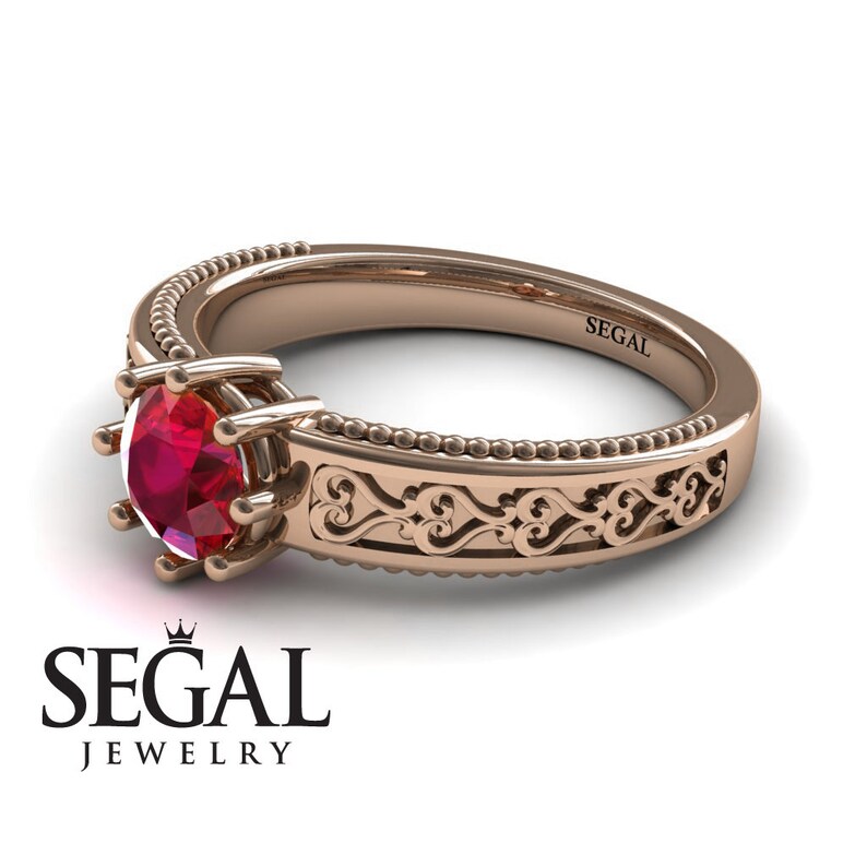 May include: A rose gold engagement ring with a red ruby gemstone and a delicate filigree design. The band is adorned with a beaded edge. The ring is engraved with the word "SEGAL".
